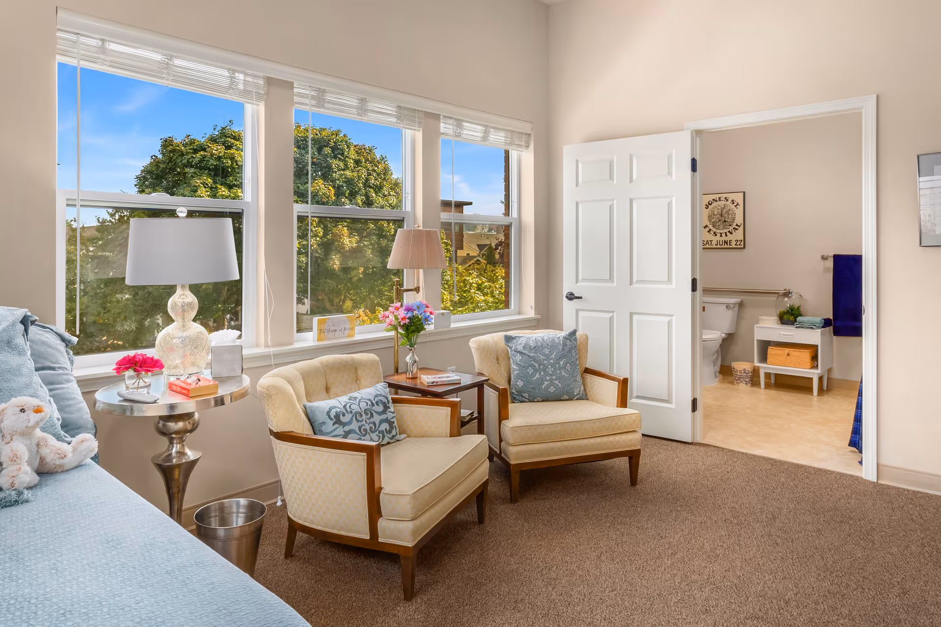 A bright and cozy living room area with two beige armchairs featuring blue patterned pillows, a side table with a lamp and flowers, and a blue sofa with a stuffed animal on it. Large windows show green trees outside. An open door reveals a bathroom with a toilet, towel rack, and small cabinet.