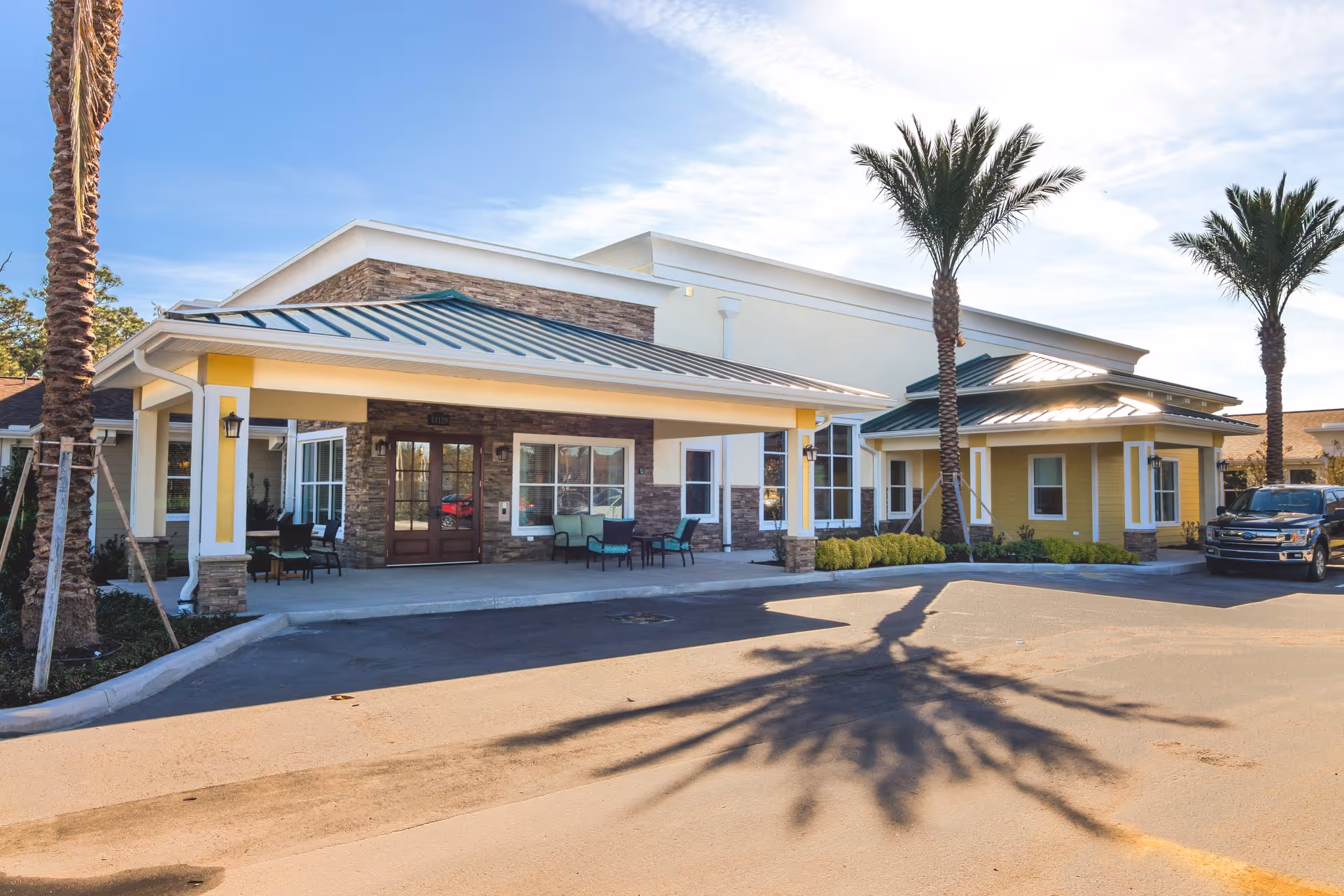 Exterior view of CERTUS Premier Memory Care Living facility showing the entrance with a covered drop-off area, stone and yellow siding, palm trees, outdoor seating, and a parked black truck.