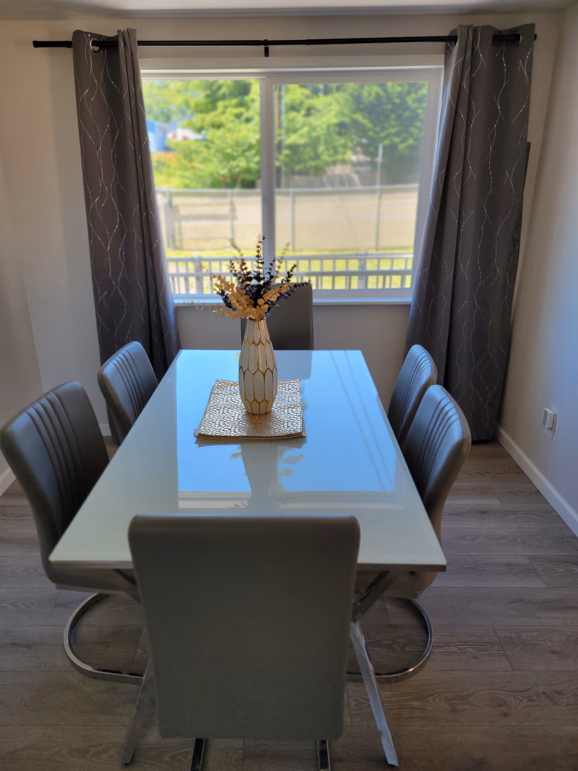 A dining room with a rectangular glass table surrounded by six gray chairs. A decorative vase with flowers is placed on a golden placemat in the center of the table. Behind the table is a large window with gray curtains, showing greenery outside.