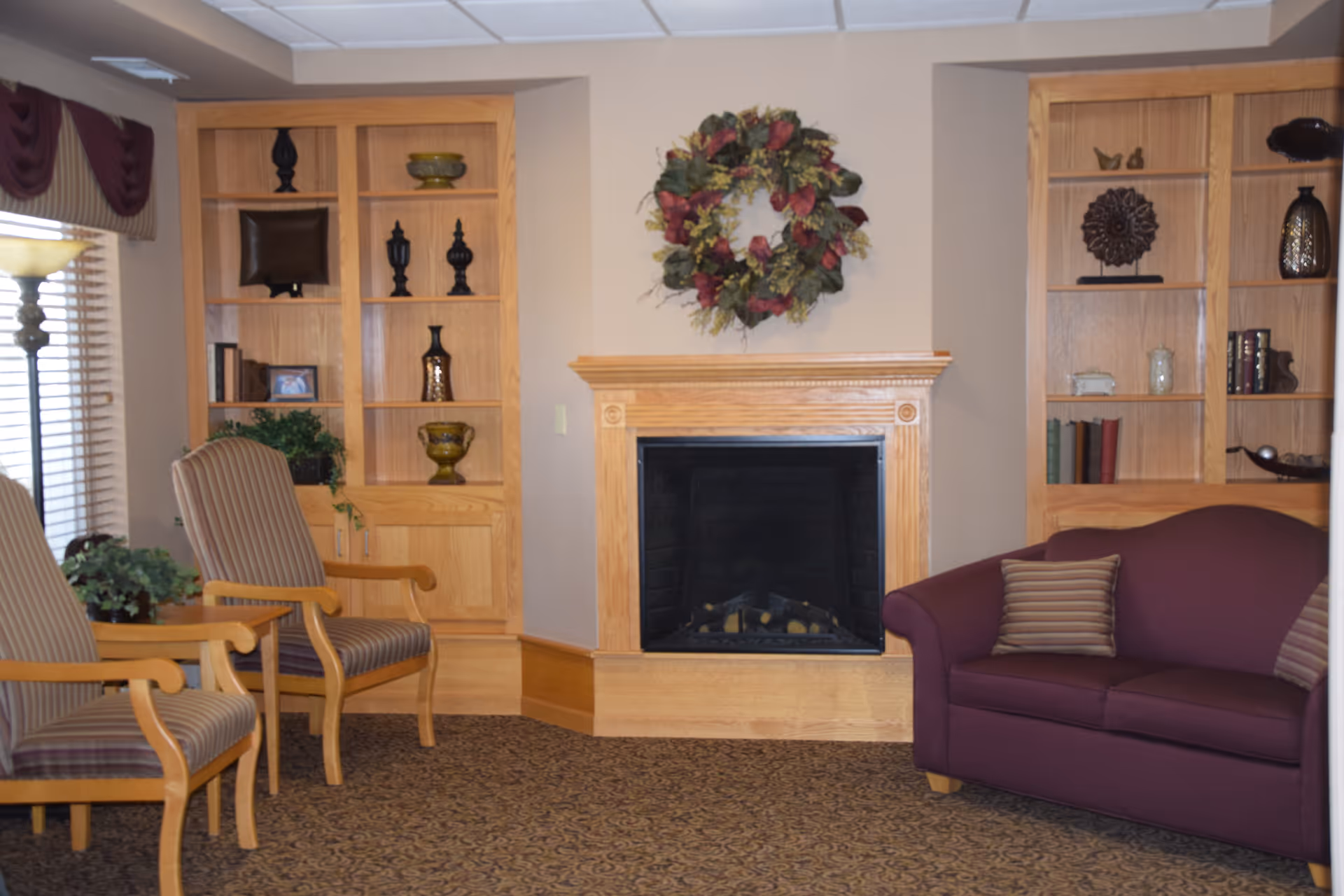 A cozy living room area with a wooden fireplace in the center, decorated with a floral wreath above it. On either side of the fireplace are built-in wooden shelves holding decorative items and books. To the left, there are two striped armchairs, and to the right, a maroon loveseat with striped pillows. A floor lamp and window with blinds and valance are visible on the left side.