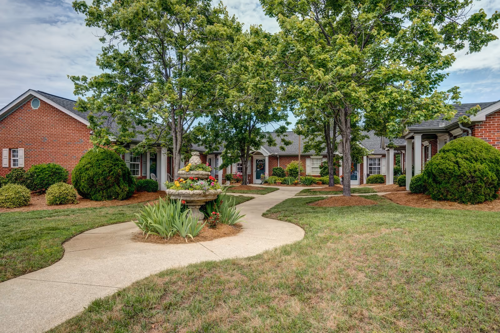 Landscaped courtyard in front of a brick retirement building with a curved walkway, trees, and a tiered flower fountain.
