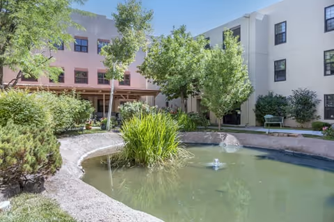 Outdoor courtyard area at Brookdale Santa Fe featuring a small pond with a fountain, surrounded by greenery including trees and bushes, with a three-story building in the background.