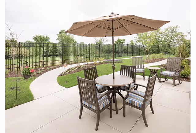 Outdoor patio area with a round table, four chairs with striped cushions, two additional chairs, a small side table, and a large beige umbrella. The patio is surrounded by a curved walkway, green grass, plants, and a black metal fence with trees in the background.