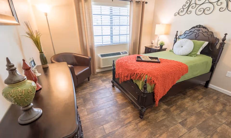 A cozy bedroom with a single dark wooden bed featuring a green bedspread and a red throw blanket. There is a round white pillow and a green pillow on the bed. A tray with a book and a cup is placed on the bed. Next to the bed is a dark wooden nightstand with a lamp and a small plant. A brown leather armchair is positioned near a window with beige curtains and blinds. A dark wooden dresser with decorative items and a tall floor lamp are also visible. The floor has a wood-like finish.