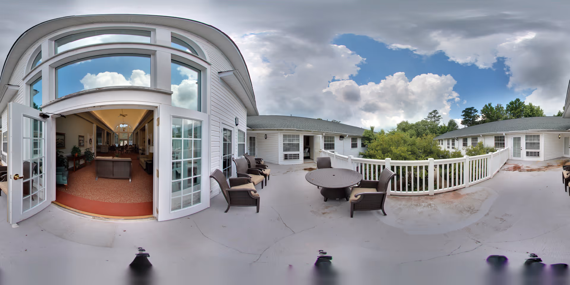 Outdoor patio area at Oaks at Tucker with several chairs and a round table. The patio is surrounded by white railings and overlooks greenery. Large glass doors open into an interior common area with seating and warm lighting.