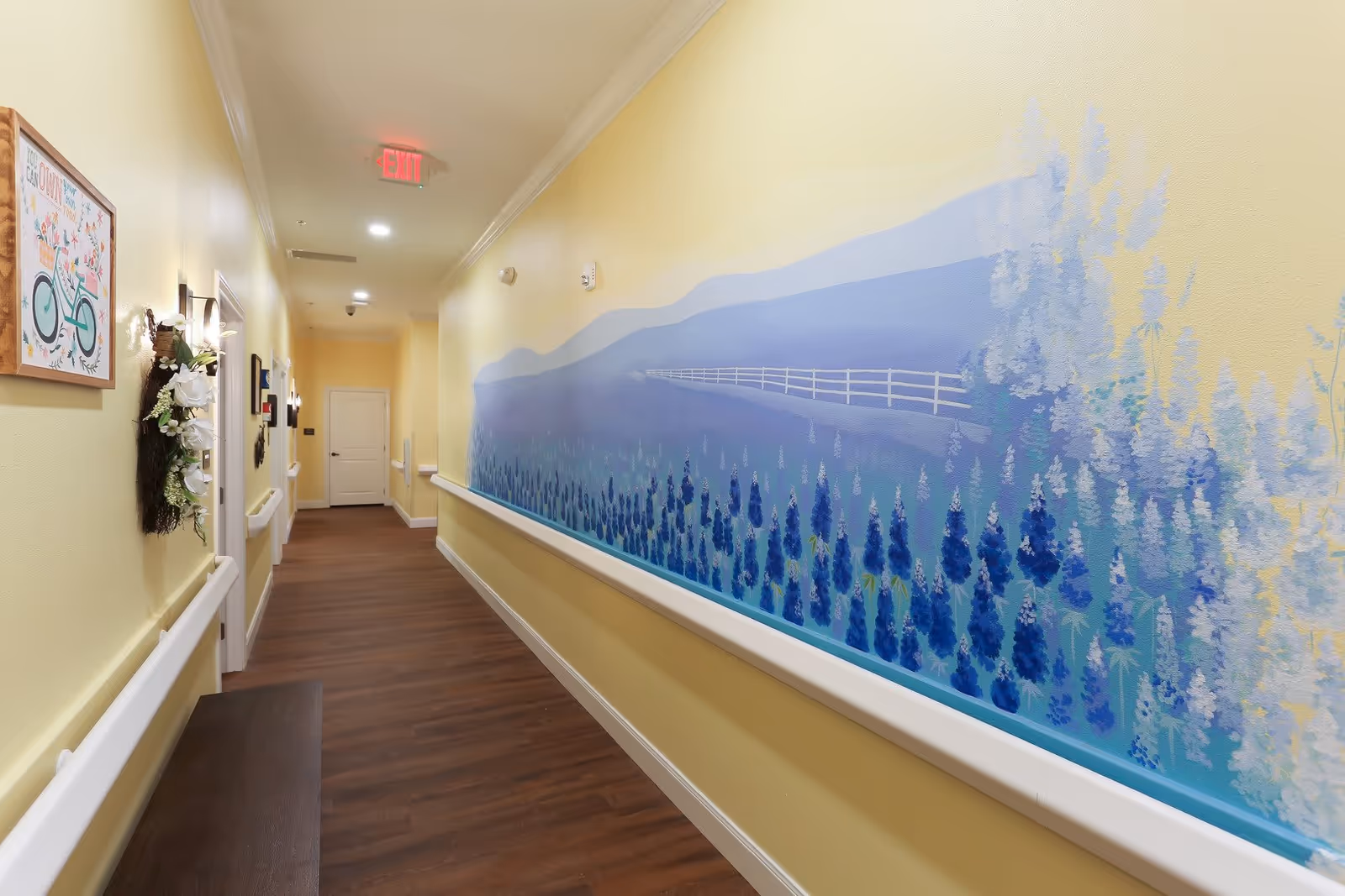A well-lit hallway with yellow walls and wooden flooring in Serenity Oaks Assisted Living and Memory Care. The right wall features a large mural of a blue and purple floral landscape with a white fence and hills in the background. The left wall has framed artwork and a decorative floral wreath. There are handrails on both sides of the hallway and a white door at the end. An illuminated exit sign is visible on the ceiling.