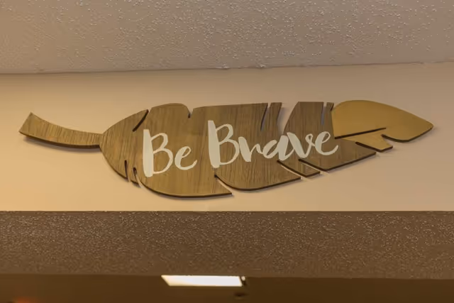 A wooden decorative sign shaped like a feather with the words 'Be Brave' written on it in white letters, mounted on a beige wall above a doorway.