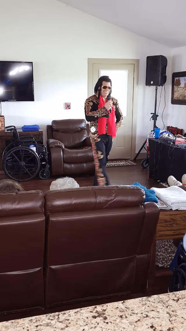 An Elvis Presley impersonator wearing a black and gold jumpsuit with a red scarf is performing with a microphone in a living room area of an assisted living facility. There are brown leather couches, a wheelchair, a TV mounted on the wall, and a speaker setup. Some elderly residents are seated on the couches watching the performance.