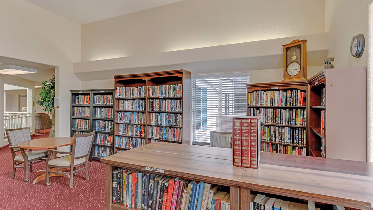 Interior view of a library or reading room in a senior living facility with multiple bookshelves filled with books, a wooden table with chairs, and a clock on top of one bookshelf. The room has carpeted flooring and a window with blinds allowing natural light.