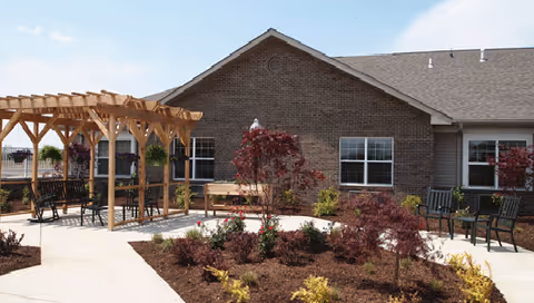 Outdoor patio area at Blair Ridge Health Campus featuring a wooden pergola with hanging flower baskets, several benches, landscaped garden beds with small trees and shrubs, and a brick building in the background under a partly cloudy sky.