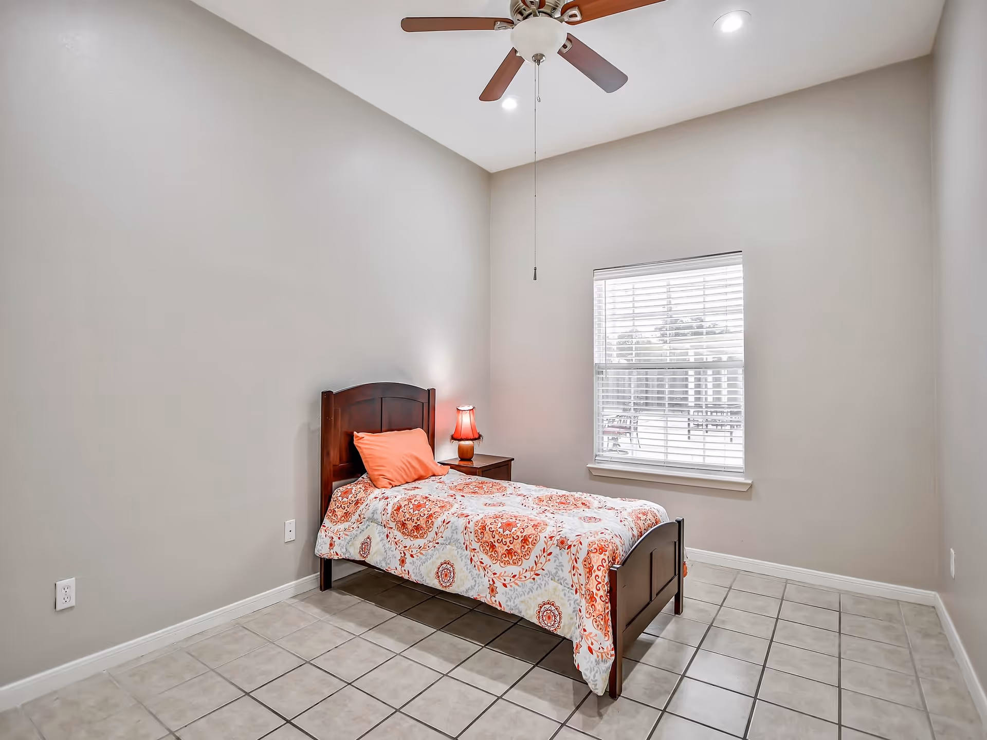 A small bedroom with a single wooden bed featuring a colorful orange and white patterned bedspread and an orange pillow. Next to the bed is a wooden nightstand with a small red lamp. The room has beige walls, a tiled floor, a window with white blinds, and a ceiling fan with wooden blades.