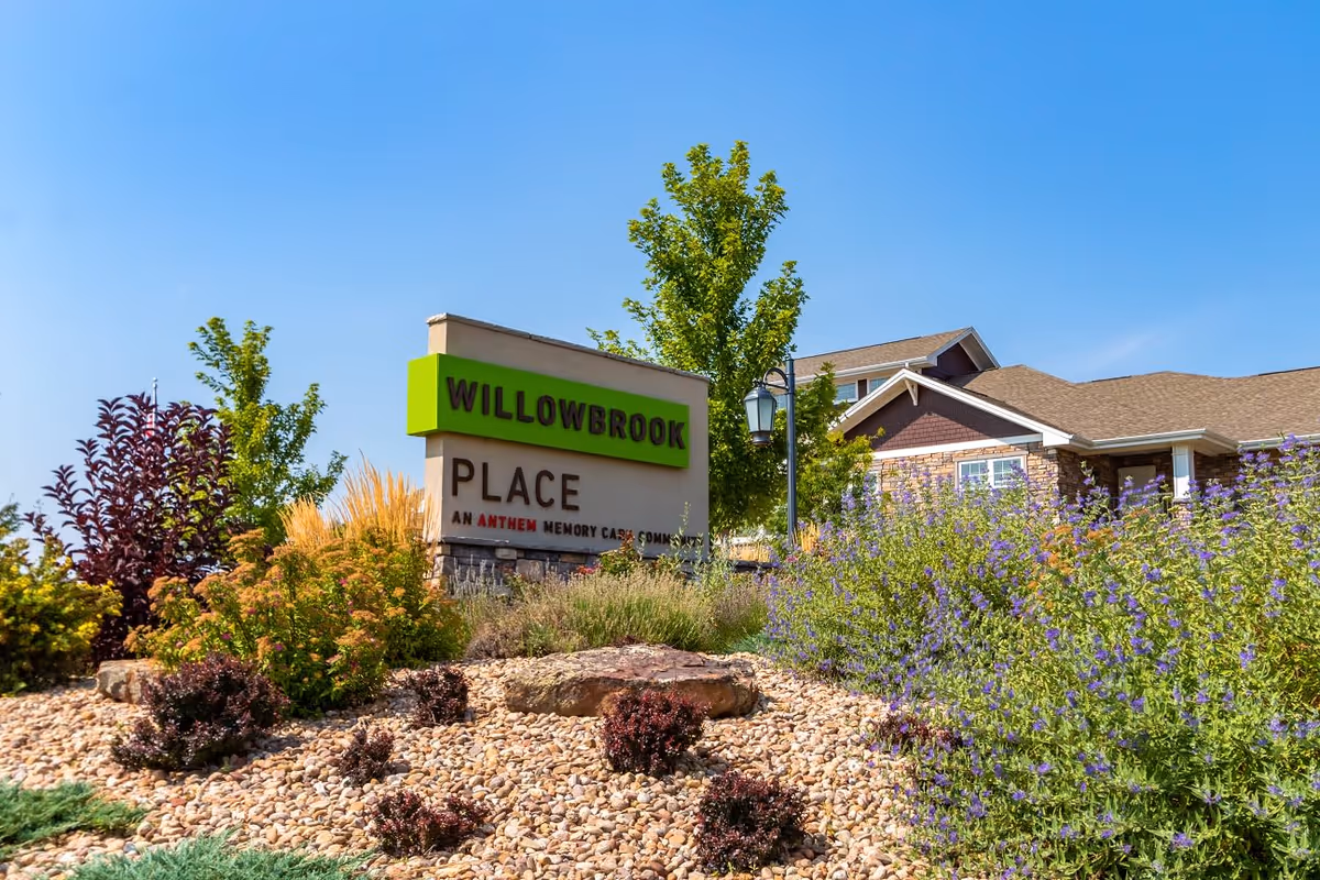 Outdoor view of the entrance sign for Willowbrook Place, an Anthem Memory Care Community, surrounded by landscaped bushes, rocks, and trees under a clear blue sky.