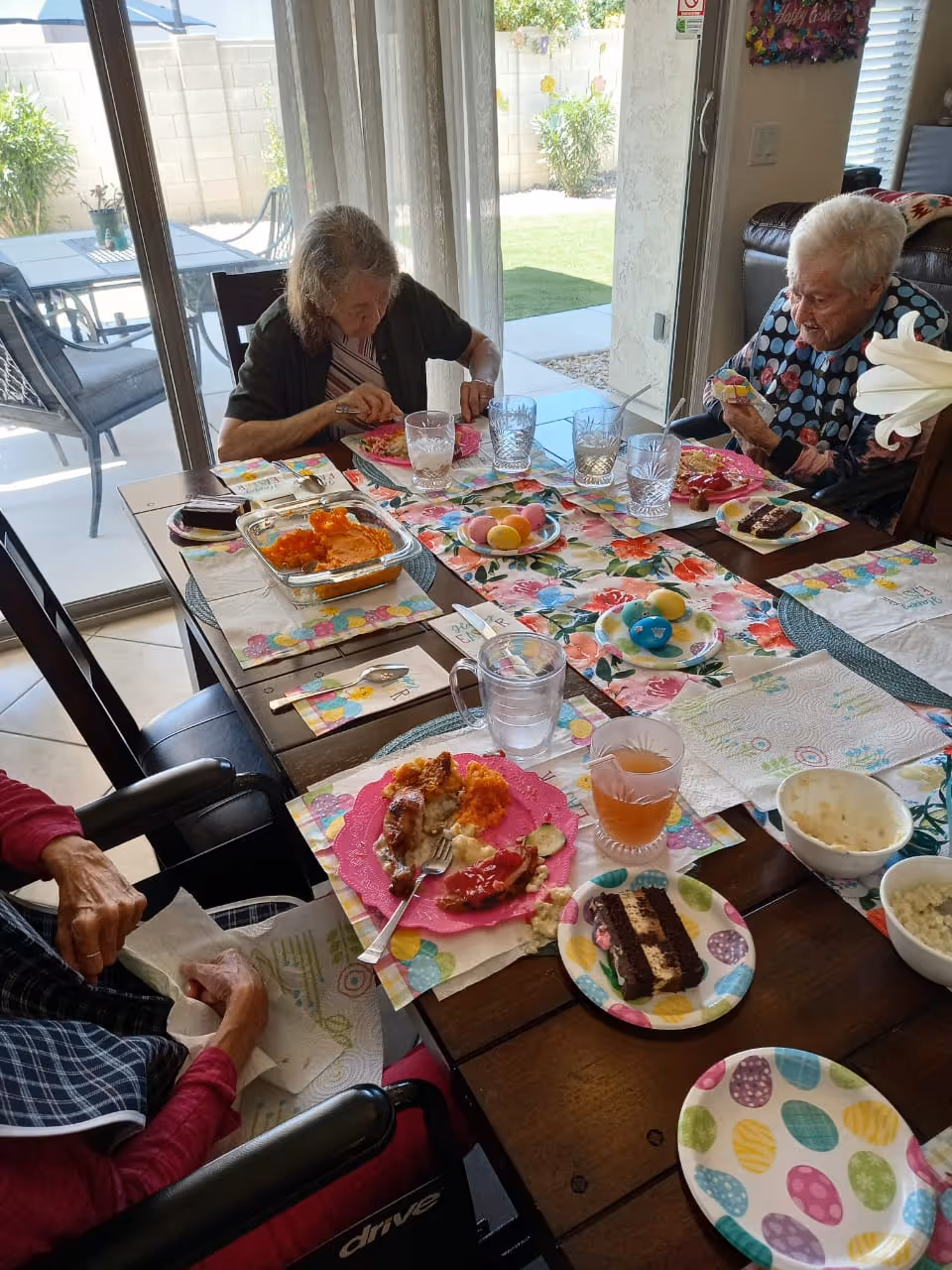 Three elderly women sitting around a dining table with colorful Easter-themed placemats and plates, eating a meal that includes meat, mashed potatoes, vegetables, and dessert. The table is set near large glass sliding doors that open to a backyard with patio furniture and greenery.