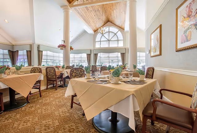 Bright dining room with neatly set tables, upholstered chairs, large arched windows and a vaulted wood ceiling with columns.