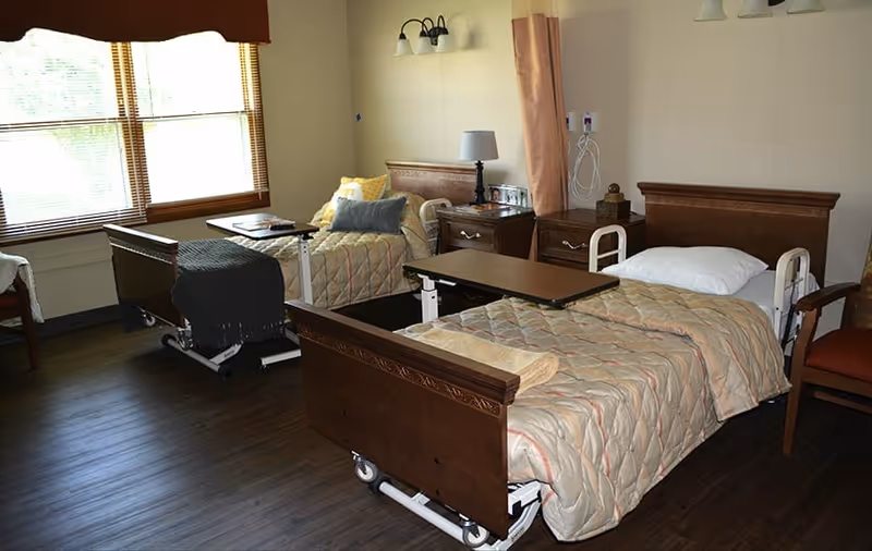 A room with two hospital-style beds with wooden headboards and footboards, each bed covered with a beige quilted blanket. There are bedside tables with lamps between the beds, a chair to the right, and large windows with blinds letting in natural light. The floor is dark wood, and the room has a calm, clean atmosphere.