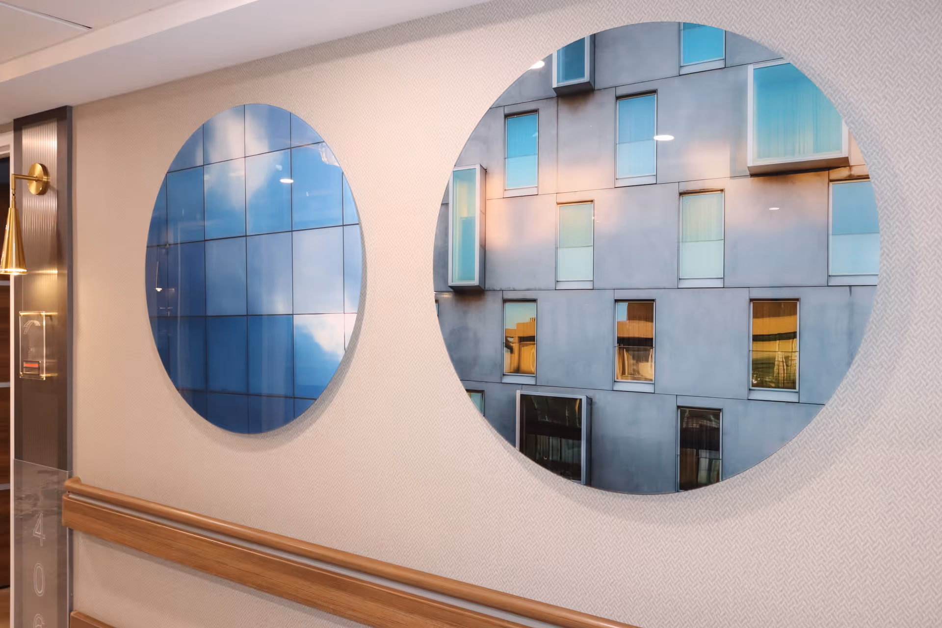Two large circular mirrors mounted on a beige textured wall in a hallway. The mirrors reflect images of building windows and a blue sky with clouds. Below the mirrors is a wooden handrail attached to the wall.