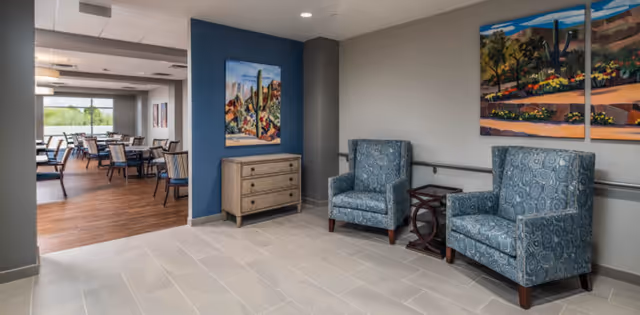 Interior view of a senior living facility showing a seating area with two patterned blue armchairs and a small wooden table between them. The walls are decorated with colorful landscape paintings. In the background, there is a dining area with multiple tables and chairs, large windows letting in natural light, and wooden flooring.