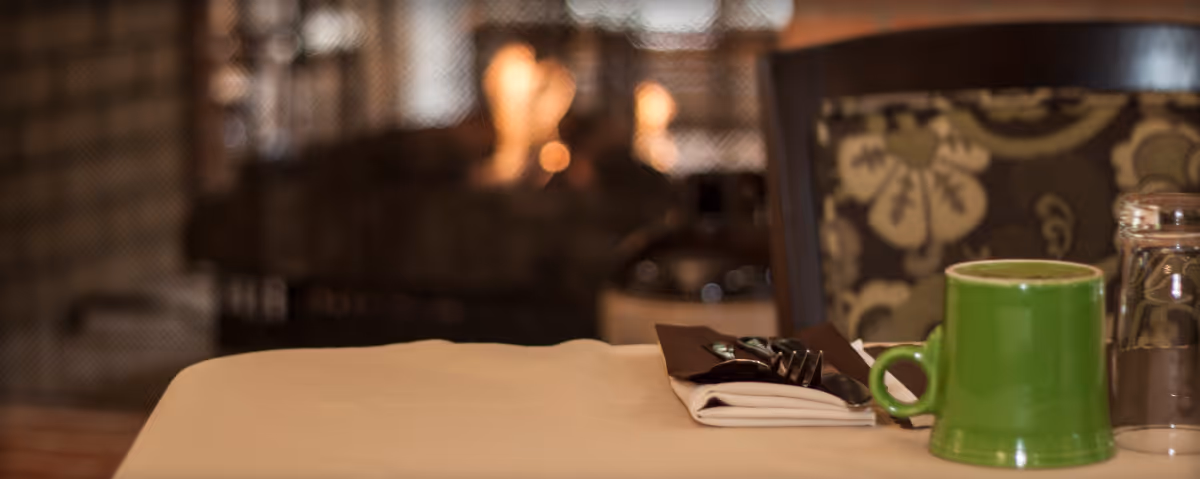 A close-up view of a dining table set with a green coffee mug, a glass, and silverware wrapped in a napkin. In the background, there is a softly glowing fireplace and a chair with a floral patterned backrest.