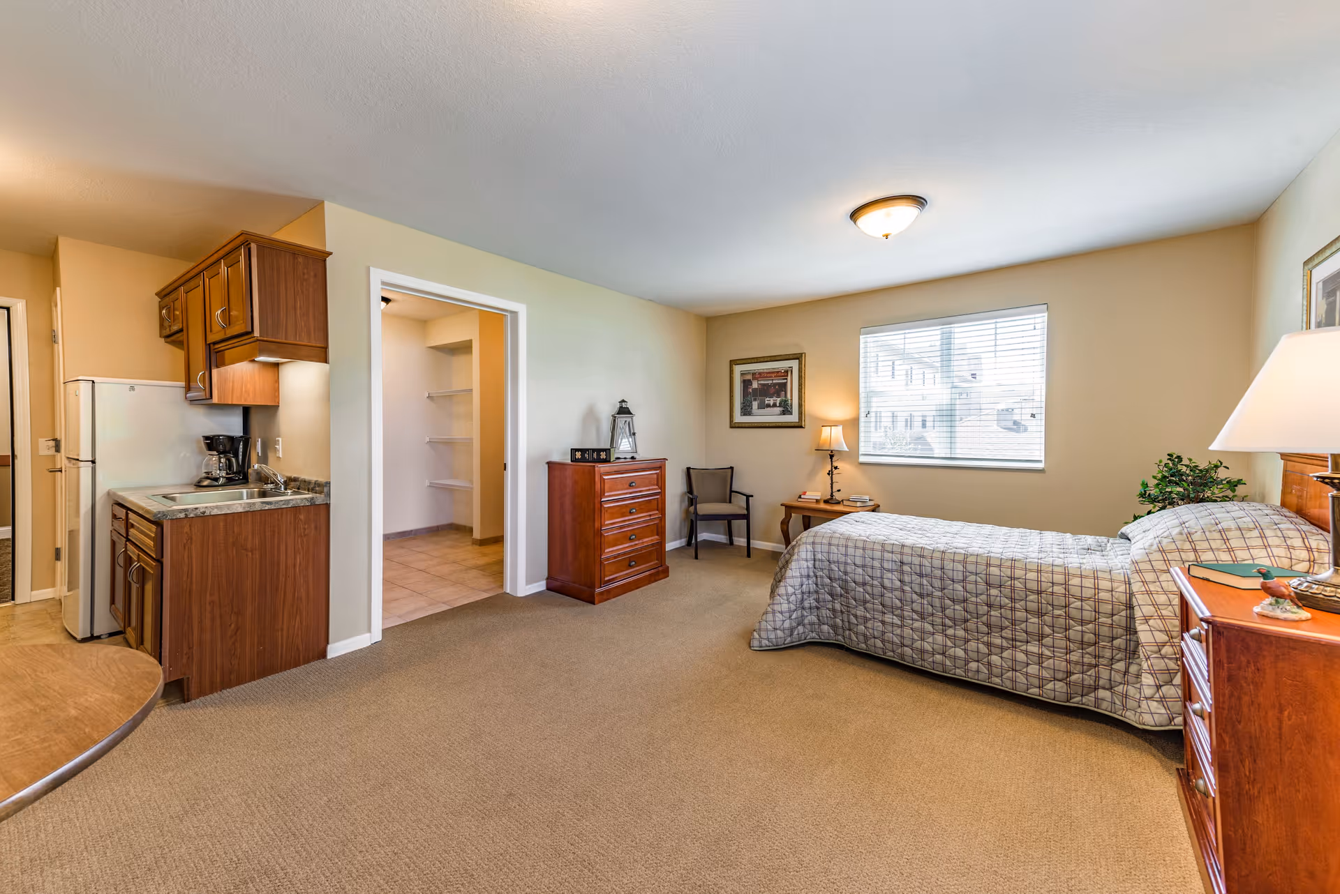 Furnished studio apartment room with a bed, dresser, chair, and small kitchenette under warm lighting.
