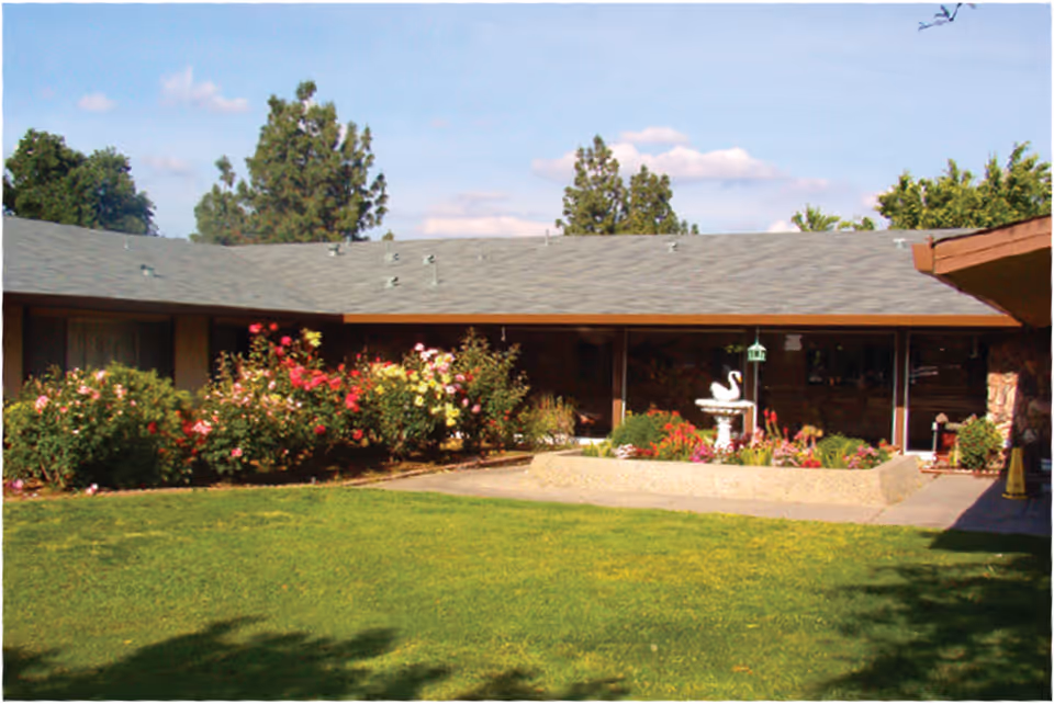 Outdoor courtyard area of a senior living facility with a green lawn, flower beds with colorful flowers, a white swan statue fountain, and a building with a gray roof and large windows in the background under a blue sky with some clouds.