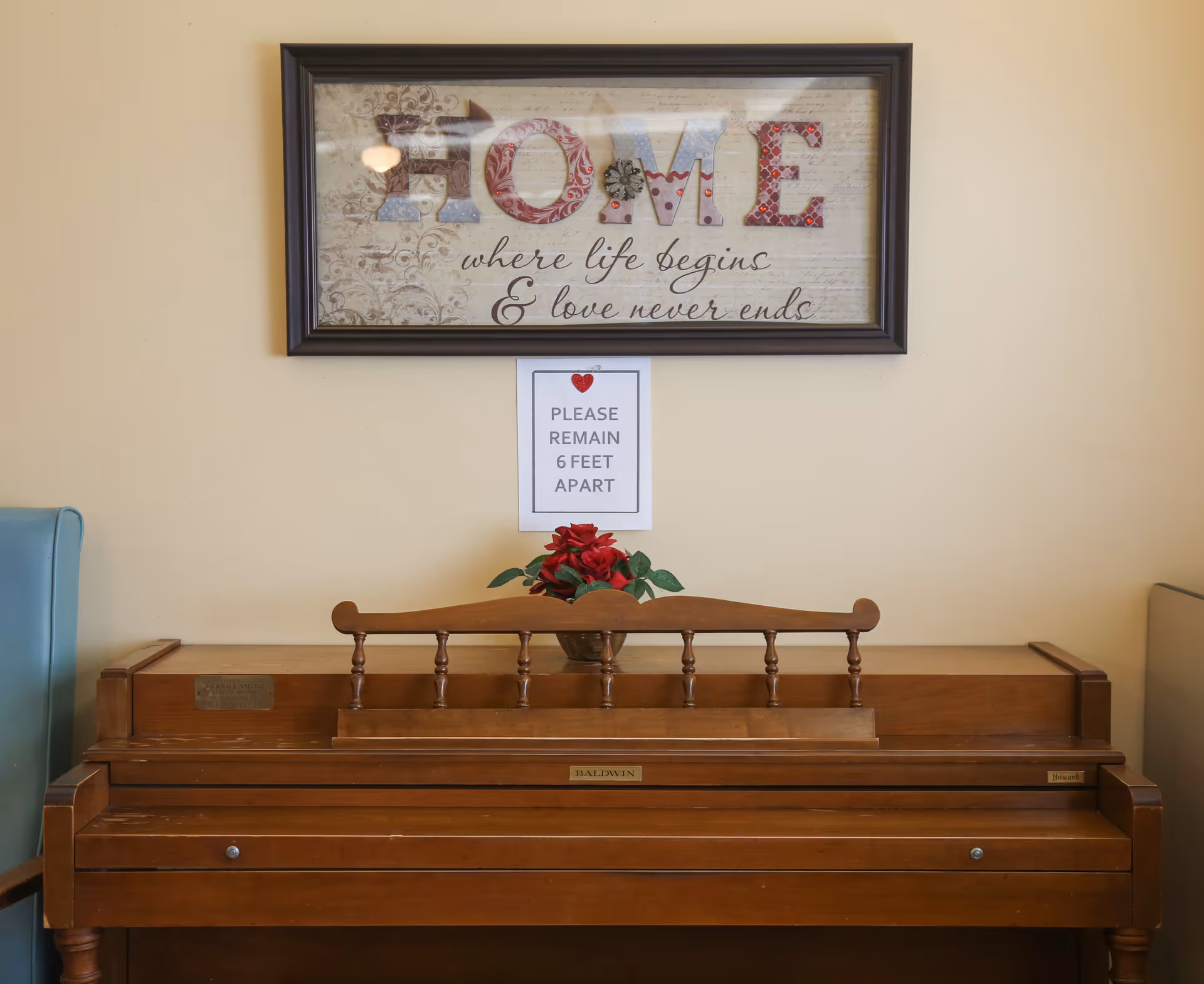 A wooden Baldwin piano with a small vase of red flowers on top, positioned against a beige wall. Above the piano is a framed decorative sign that reads 'HOME where life begins & love never ends.' Below the sign, a paper notice says 'PLEASE REMAIN 6 FEET APART.'