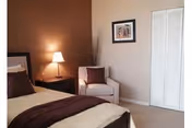 A cozy bedroom with a bed featuring beige and brown bedding, a nightstand with a lamp, a white armchair with a brown cushion, a framed picture on the wall, and a white closet door.
