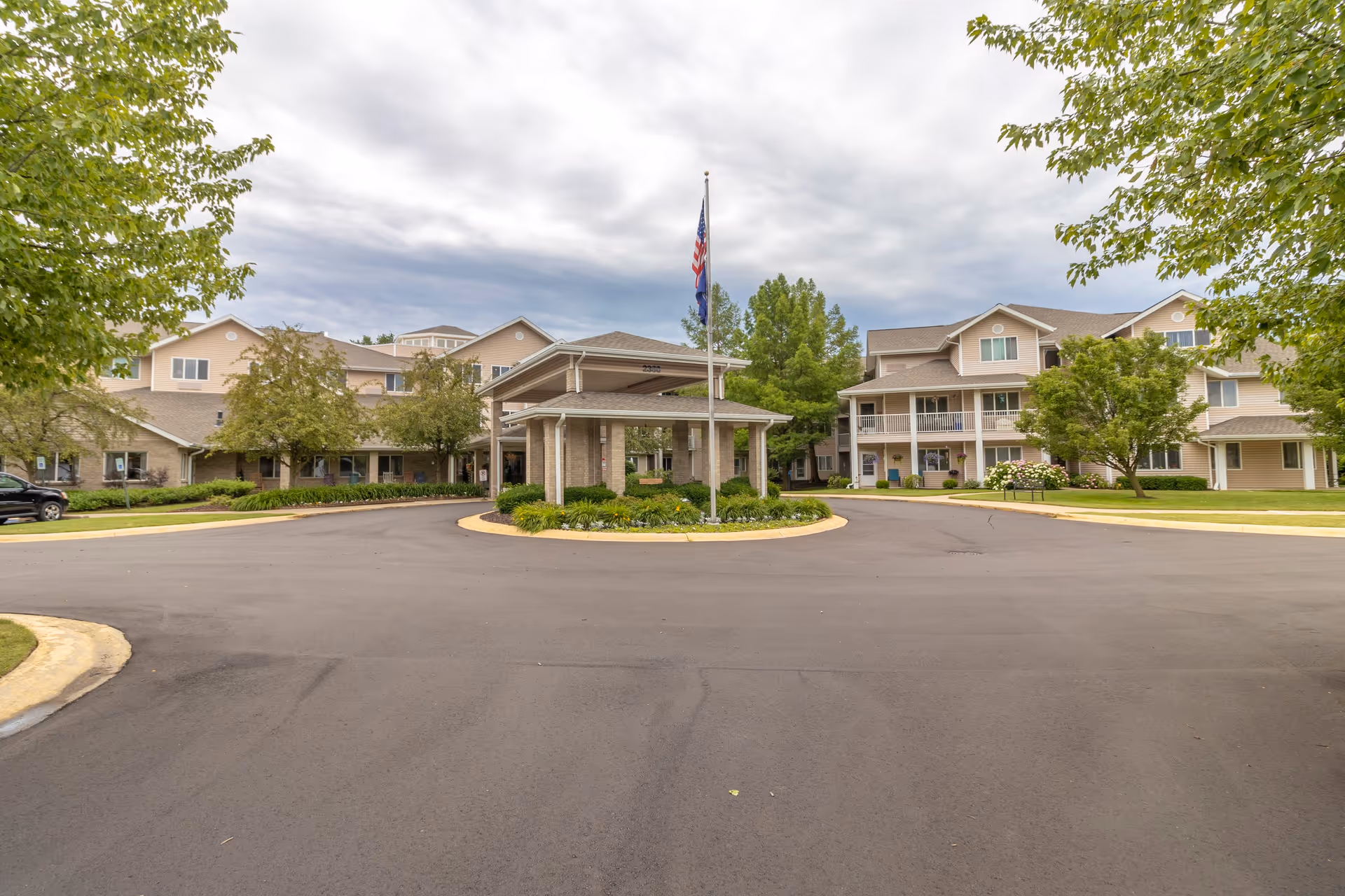 Exterior view of StoryPoint Wyoming senior living facility showing a circular driveway with a covered entrance and flagpole in the center. The building is multi-story with beige siding and surrounded by trees and greenery under a cloudy sky.