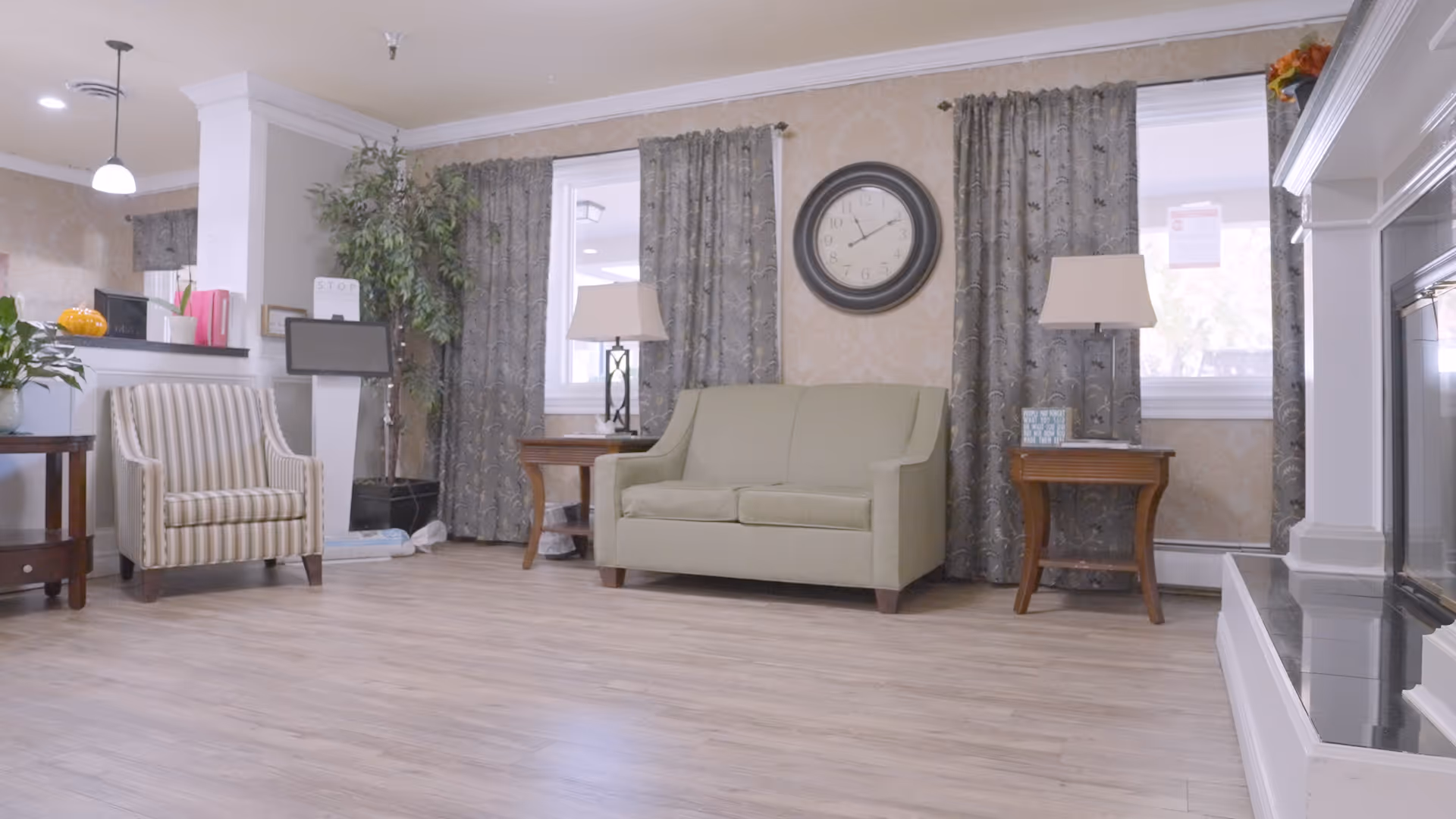 A cozy living room area with a light green loveseat centered between two wooden side tables, each with a lamp. To the left is a striped armchair next to a small table with a plant. Behind the loveseat are two windows with patterned curtains and a large round clock on the wall. The floor is light wood, and a white fireplace is visible on the right side.