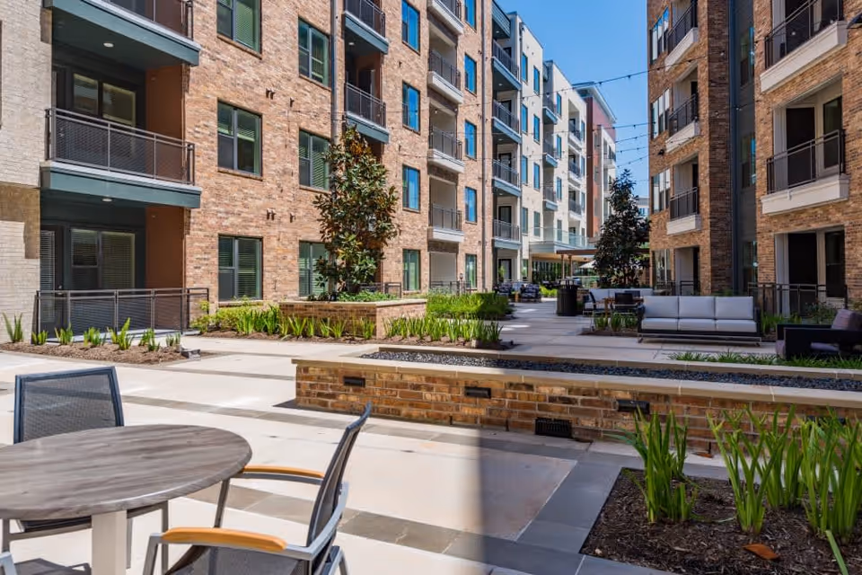 Outdoor courtyard area of a senior living facility featuring modern multi-story brick and stucco buildings with balconies. The courtyard includes seating areas with tables and chairs, planted greenery, and string lights overhead under a clear blue sky.