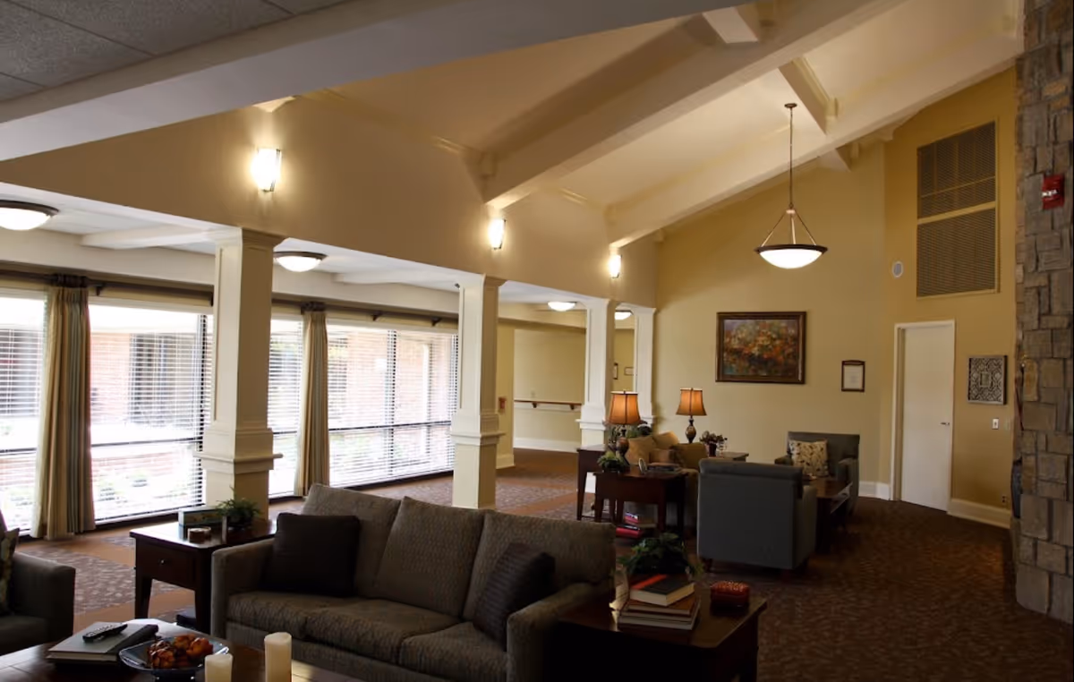 A spacious and warmly lit living room area in a senior living facility with comfortable sofas and armchairs arranged around wooden side tables. The room features large windows with blinds and curtains, a high vaulted ceiling with exposed beams, wall sconces, and a hanging light fixture. There is a stone wall on the right side and framed artwork on the walls.