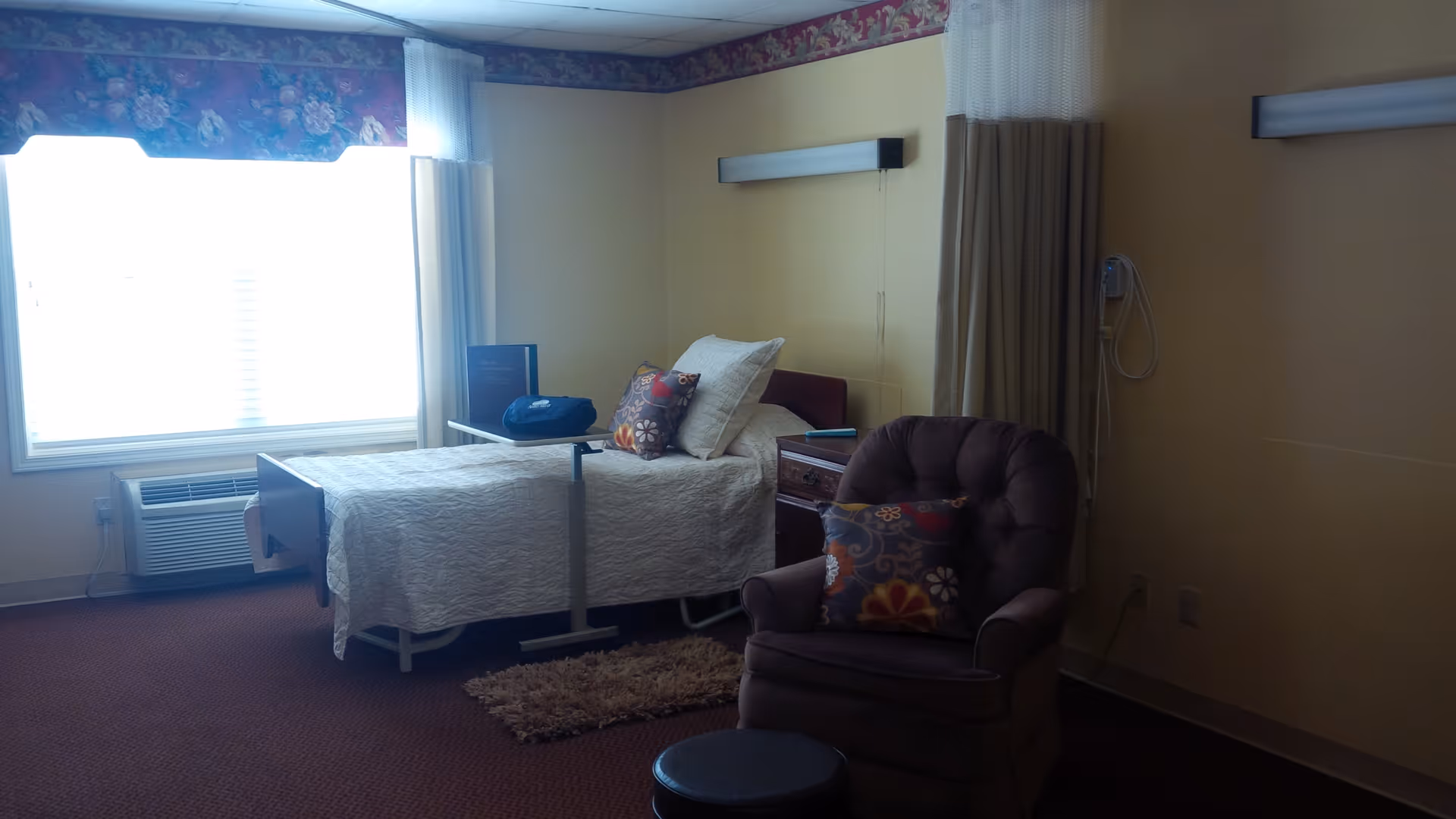 A senior living facility bedroom with a single bed covered in a white quilt, two decorative pillows, a bedside table with a phone, a brown cushioned armchair with a colorful pillow, a small round footstool, a window with white curtains and a floral valance, and a wall-mounted light fixture above the bed.