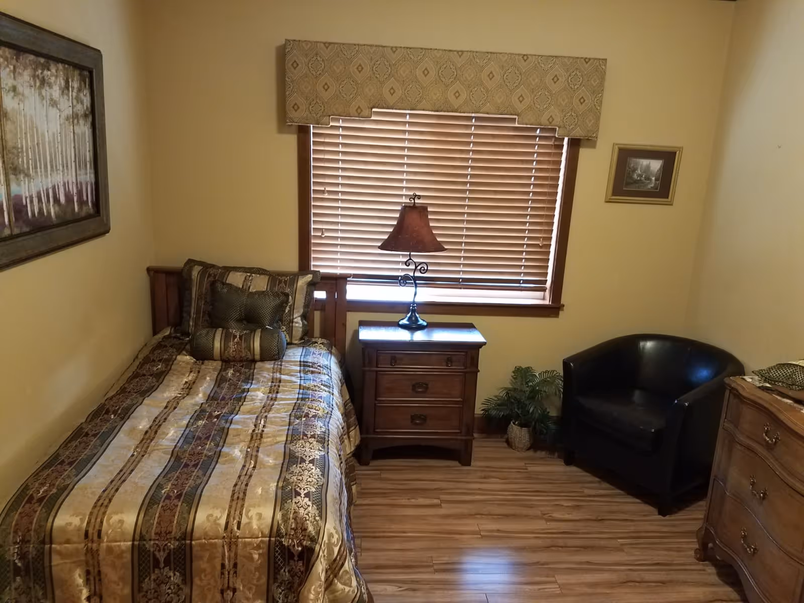 Small bedroom with a single bed, wooden nightstand and lamp, a black armchair, and a window with closed blinds.