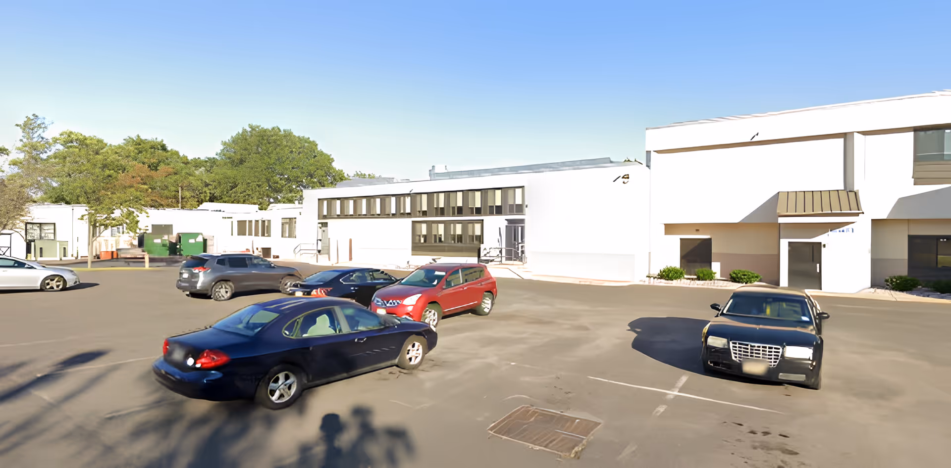 Parking lot with several cars parked in front of a white building with multiple windows and doors, surrounded by trees under a clear blue sky.