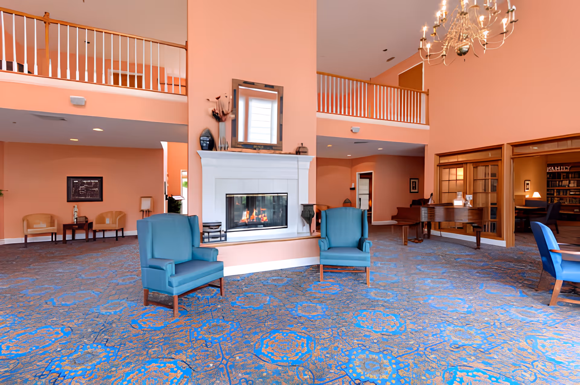 A spacious living room area with peach-colored walls and a blue patterned carpet. The room features a white fireplace with a mirror above it, two blue armchairs in front of the fireplace, a chandelier hanging from the ceiling, and a grand piano near a set of large windows. There is a balcony railing on the upper level and additional seating areas with chairs and tables in the background.