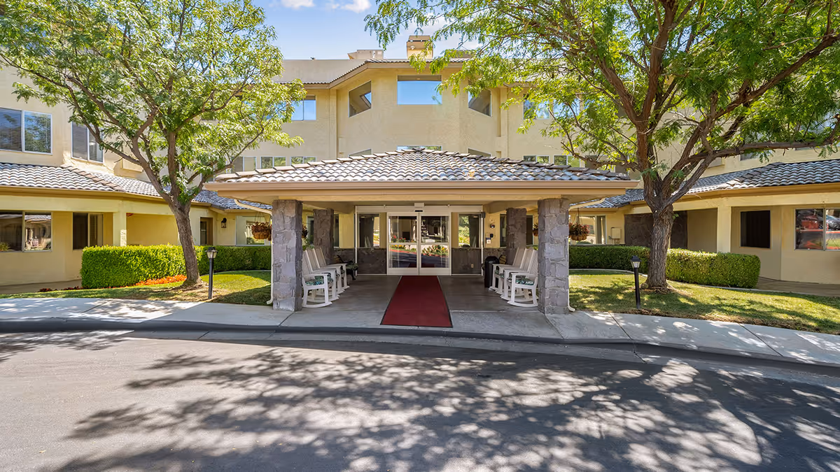 Front entrance of Columbus Estates - A Provincial Senior Living Community, featuring a covered driveway with stone pillars, rocking chairs on both sides, surrounded by green trees and bushes under a clear blue sky.