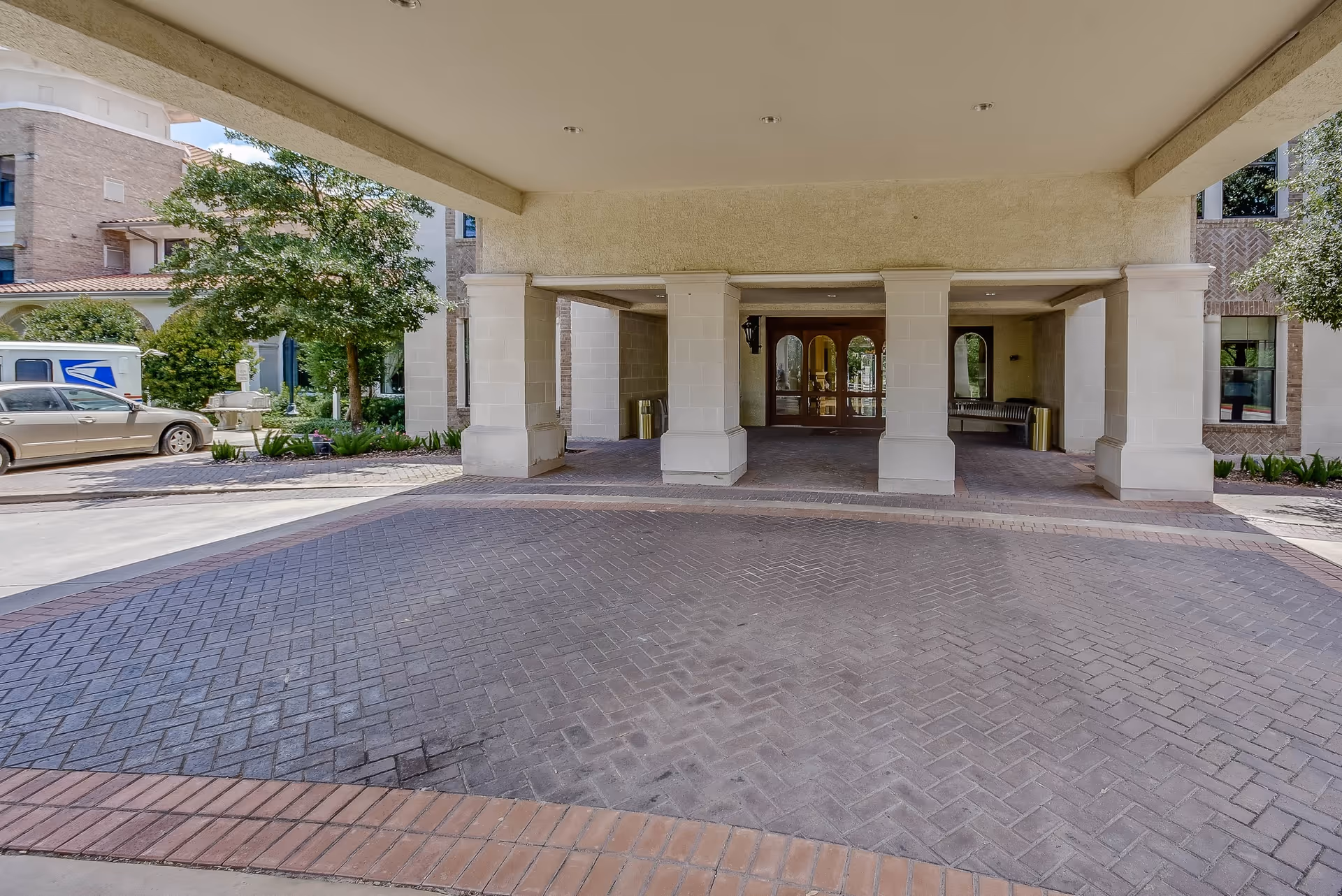 Covered driveway entrance to a building with large white pillars and double wooden doors. There are trees, a parked car, and a USPS mail truck visible on the left side outside the covered area.