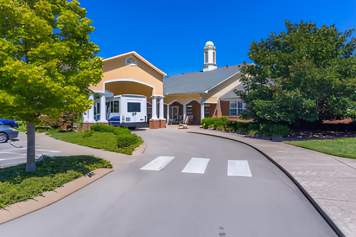 Exterior view of Charter Senior Living of Hermitage showing the driveway entrance with a covered drop-off area, surrounded by green trees and bushes under a clear blue sky.