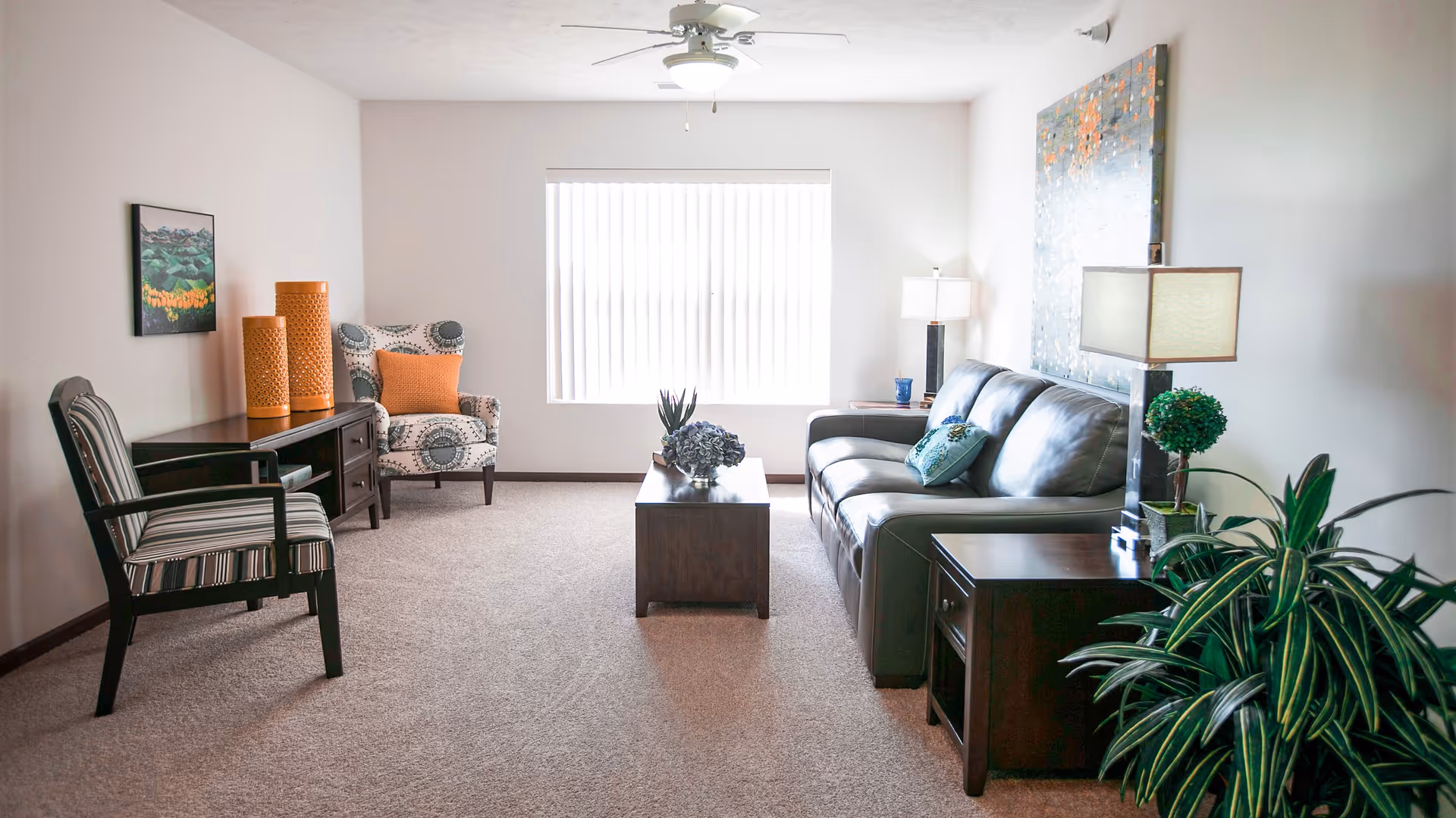 A bright living room with a large window covered by vertical blinds. The room features a dark leather sofa with decorative pillows, a wooden coffee table with a floral arrangement, two side tables with lamps, a patterned armchair with an orange pillow, a striped chair, and a wooden console table with two large orange vases. There is a ceiling fan with a light fixture and a large piece of artwork on the wall above the sofa. A green potted plant is visible in the foreground.