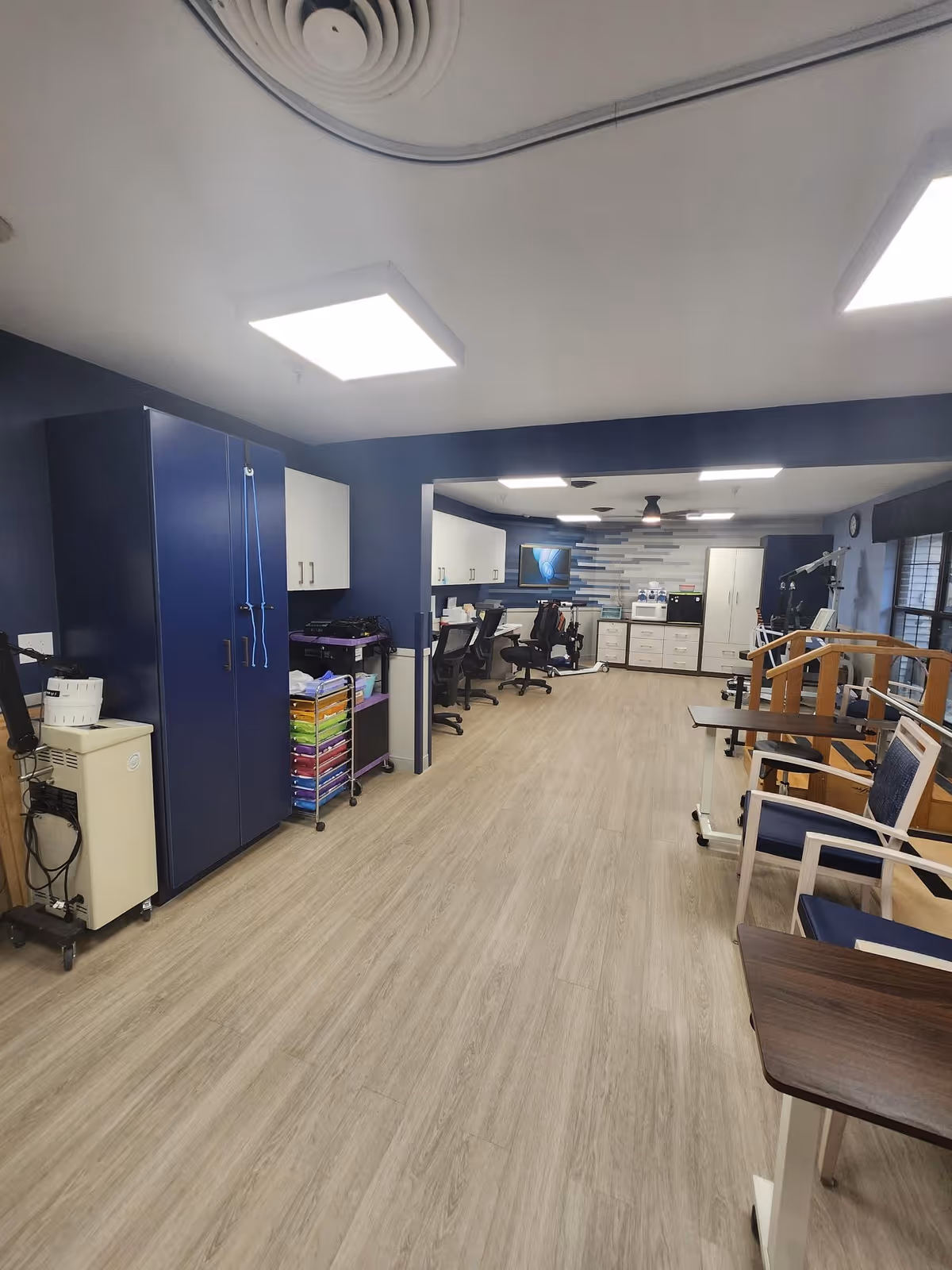 Interior view of a rehabilitation center room with wooden flooring, blue walls, and ceiling lights. The room contains office chairs, desks, cabinets, and various rehabilitation equipment including parallel bars and exercise machines. There are windows on the right side letting in natural light.