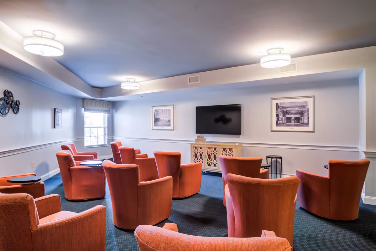 A cozy common room with multiple orange upholstered armchairs arranged in rows facing a wall-mounted flat-screen TV. The room has soft lighting from ceiling fixtures, framed black and white photographs on the walls, a window with a patterned valance, and a decorative wooden cabinet beneath the TV.