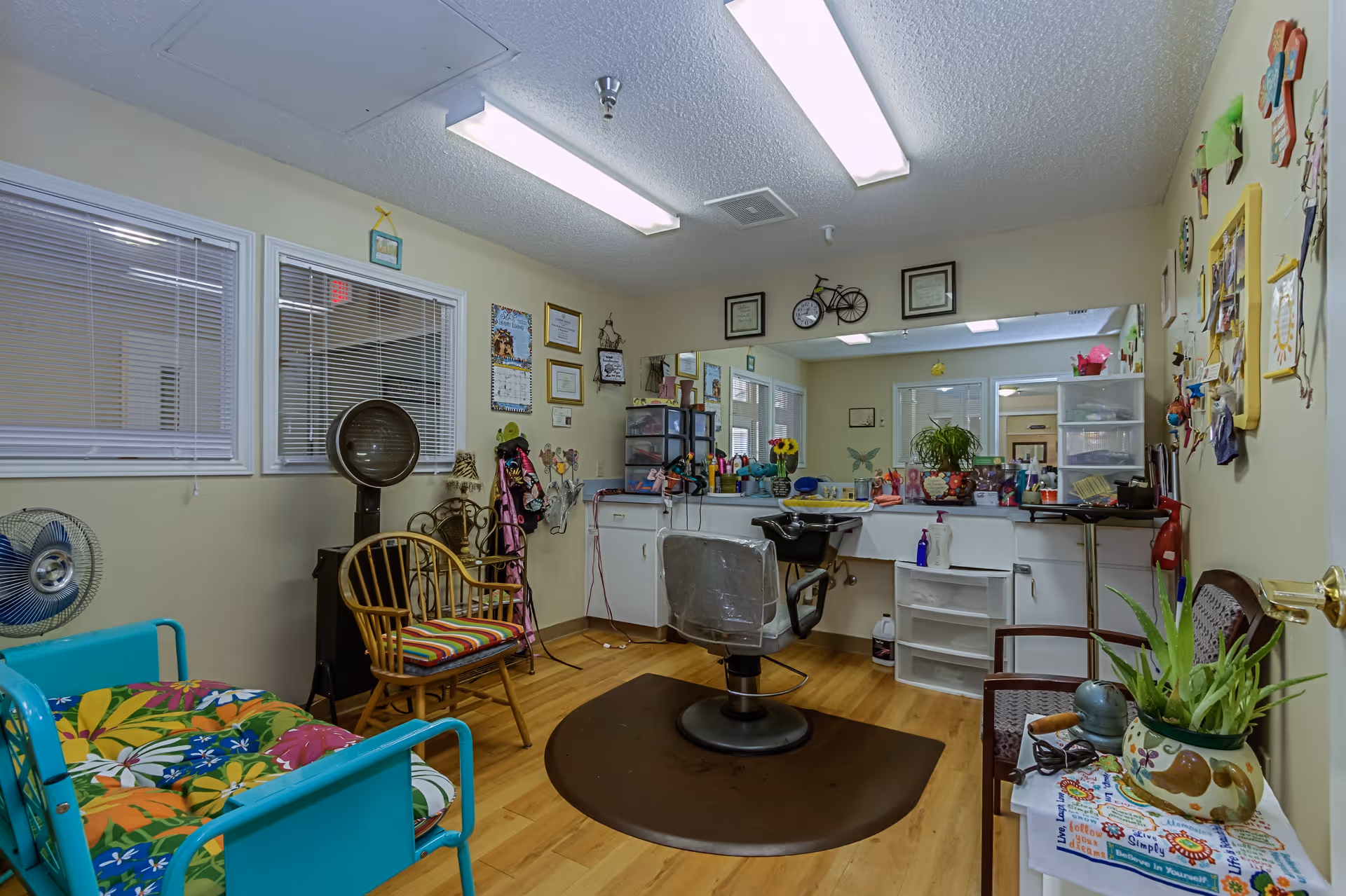 Interior of a senior living facility's hair salon room featuring a central salon chair with a protective cover, a hair washing station, a large mirror on the wall, various hair care products on the counter, colorful chairs with floral cushions, a vintage hair dryer, framed certificates and decorations on the walls, and a wooden floor.