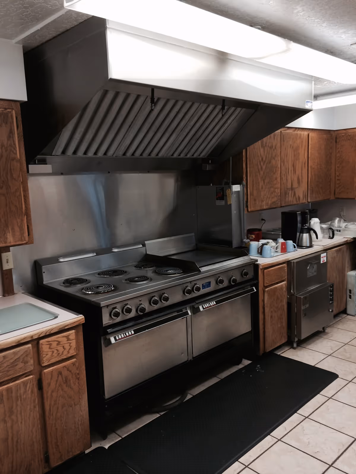 Commercial kitchen area with a large stainless steel range and hood, wooden cabinets, and countertop appliances.