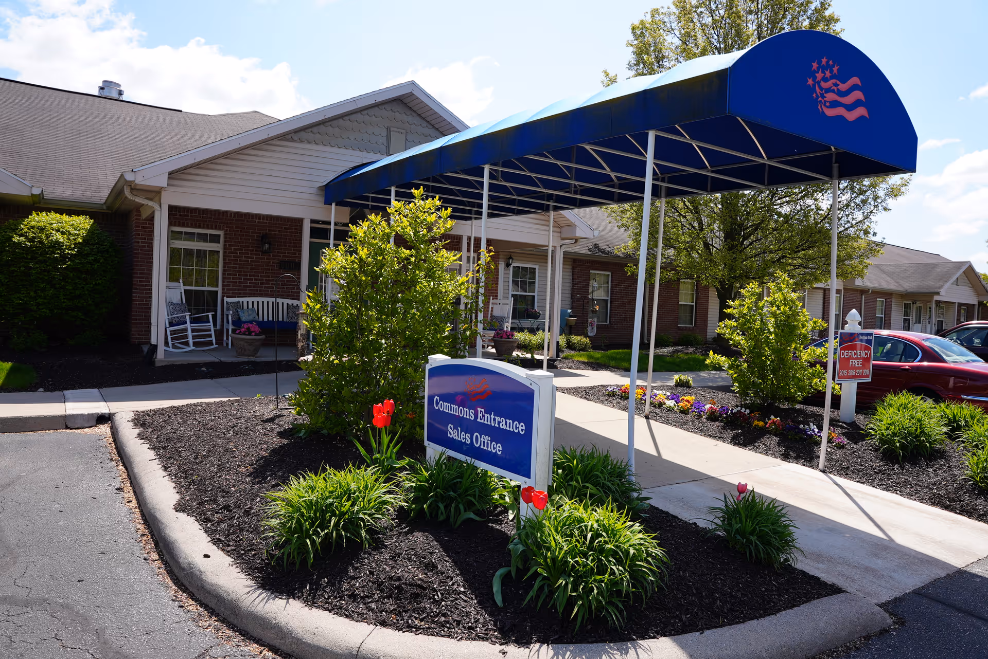 Exterior view of a senior living facility entrance with a blue canopy labeled with a red emblem. The entrance has a sign that reads 'Commons Entrance Sales Office' surrounded by landscaped plants and flowers. There are brick buildings and parked cars visible in the background under a partly cloudy sky.