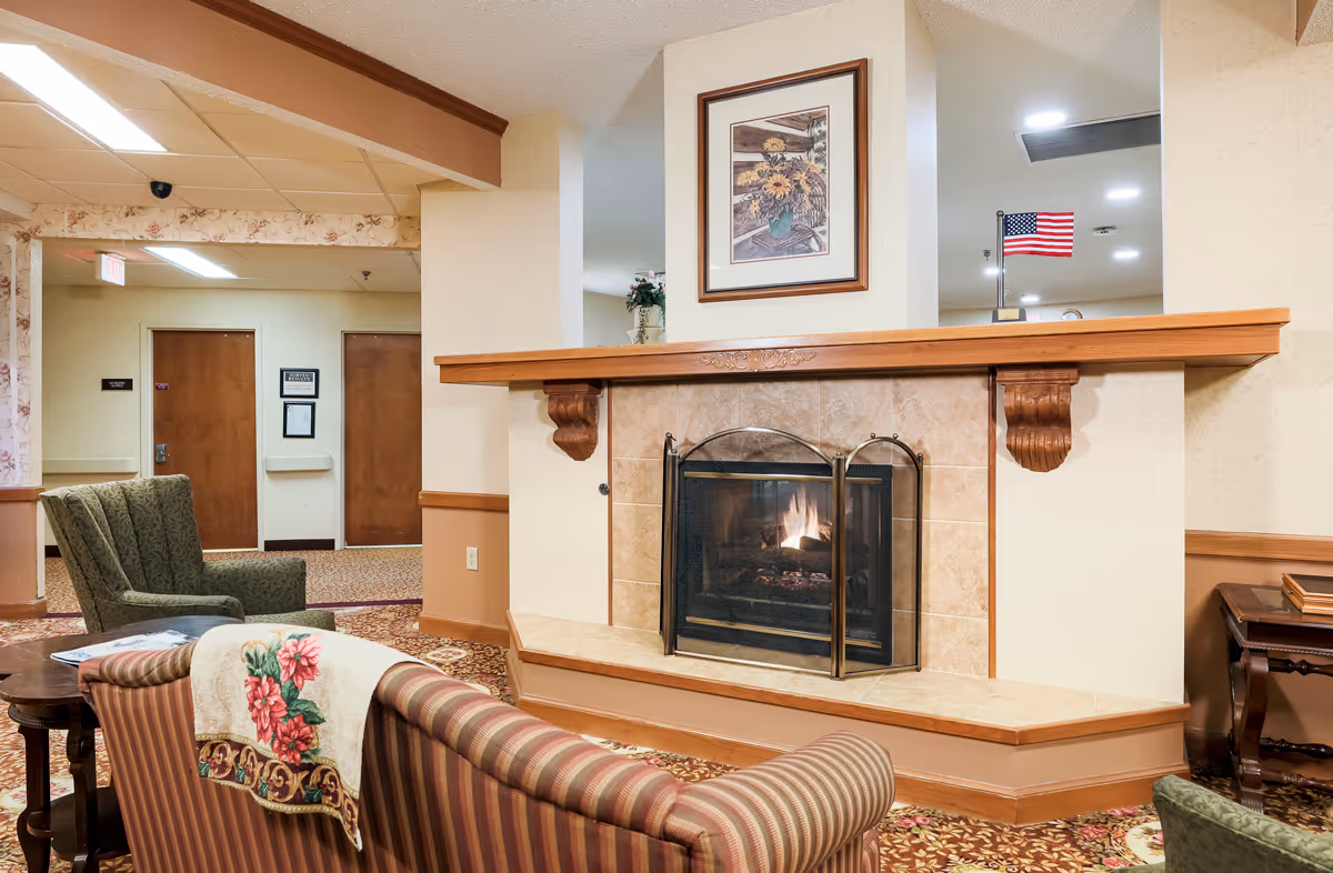 Communal lounge with a lit fireplace, striped sofa, upholstered chairs, and framed art above the mantle.