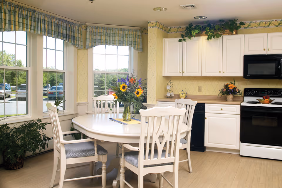 Bright kitchen and dining area with white cabinetry, black appliances, and a white dining table surrounded by four chairs. A vase with sunflowers and other flowers is placed on the table. Large windows with plaid valances let in natural light, and green plants are positioned near the windows and on top of the cabinets.