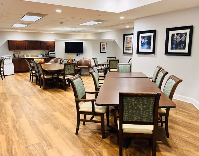 A spacious dining area with wooden floors and multiple tables surrounded by chairs. The room features a kitchenette with dark wood cabinets, a microwave, and a refrigerator. There is a wall-mounted TV and framed artwork on the walls, creating a comfortable and inviting atmosphere.