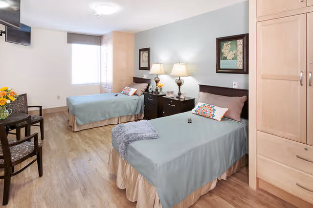 A bright and clean bedroom with two single beds covered in light blue bedspreads and beige skirts. Each bed has a decorative pillow and a remote control on top. Between the beds is a dark wooden nightstand with two lamps, flowers, and framed pictures on the wall above. The room has light wood flooring, a window with blinds, a wall-mounted TV, two chairs with a small round table holding a vase of flowers, and built-in wooden cabinetry.