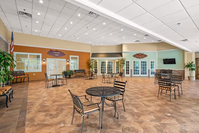 Spacious indoor seating area with multiple round tables and chairs arranged on a tiled floor. The walls are painted in warm tones with decorative signs reading 'Courtyard Café' and 'The Addison Café'. There are several potted plants and benches along the walls, and an American flag is visible in the corner. The ceiling has recessed lighting and air vents.