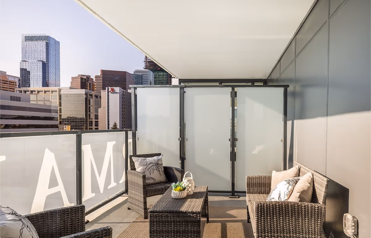 A modern outdoor balcony seating area with wicker furniture including a loveseat, two chairs, and a coffee table with decorative cushions and a small plant. The balcony has frosted glass panels and overlooks a cityscape with tall buildings under a clear sky.