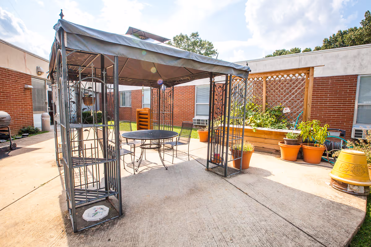 Outdoor patio area with a metal gazebo structure covering a round table and four chairs. Surrounding the patio are brick walls with windows, potted plants, a raised garden bed with greenery, and a barbecue grill on the left side. The sky is partly cloudy and the area is well-lit by sunlight.